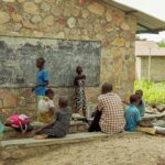 Children learning in an outdoor classroom setting with a chalkboard and stone wall backdrop.