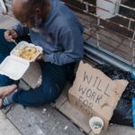 African American man sitting on a sidewalk, eating with a "Will Work for Food" sign.